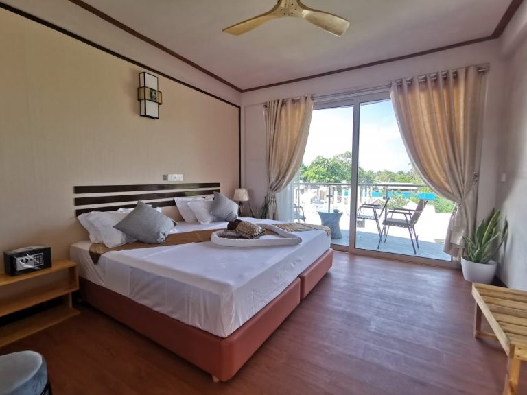 Double bed with towel heart in hotel room and view of palm trees through glass door