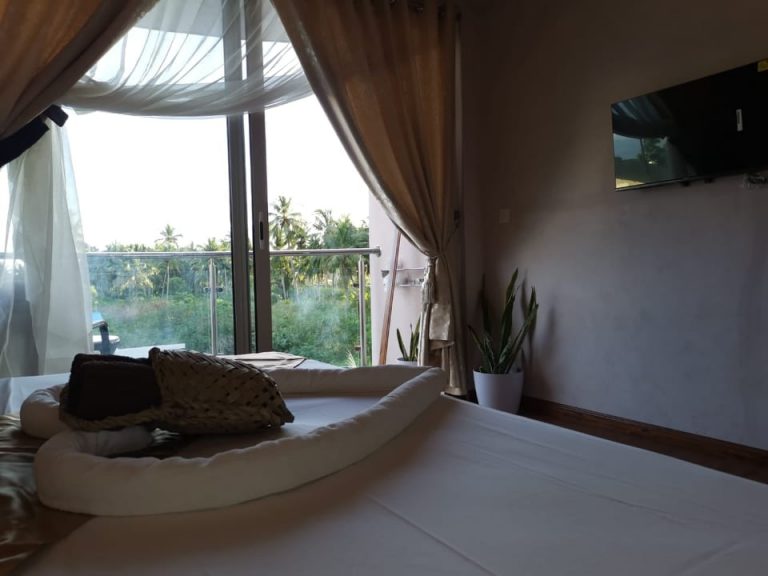 A heart made of a towel on the bed and a view through the glass door to the outdoor greenery with palm trees